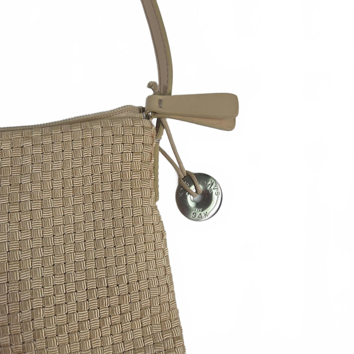 Close-up of a woven beige handbag with a leather strap and a round silver metal charm engraved with the brand name 'SOL'S'.