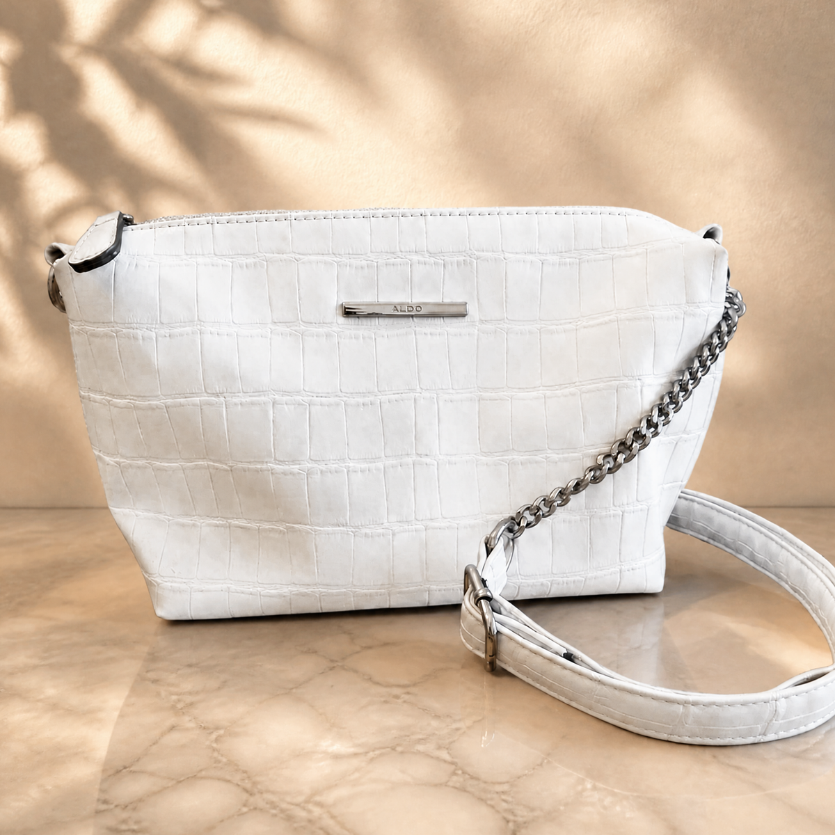 A white crocodile-embossed leather shoulder bag with a silver chain strap and a small silver metal logo plate, displayed on a beige marble surface with soft leaf shadows.