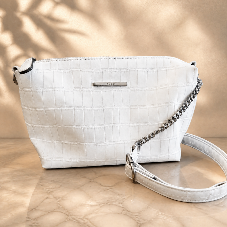 A white crocodile-embossed leather shoulder bag with a silver chain strap and a small silver metal logo plate, displayed on a beige marble surface with soft leaf shadows.